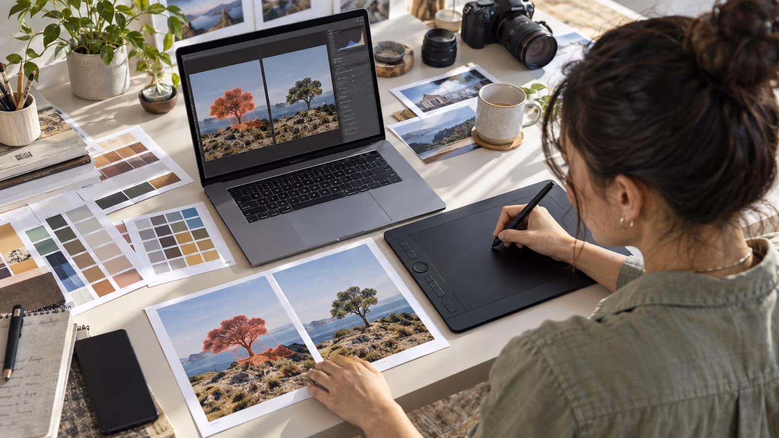 Designer using a pen tablet to mask a tree while comparing two landscape photo proofs beside a laptop and color swatches